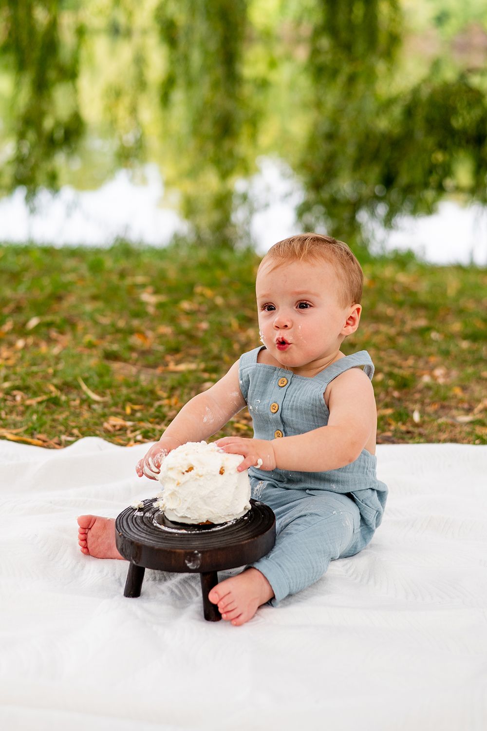 Baby in denim overalls sits on white blanket with cake on wooden stand during outdoor photo session.