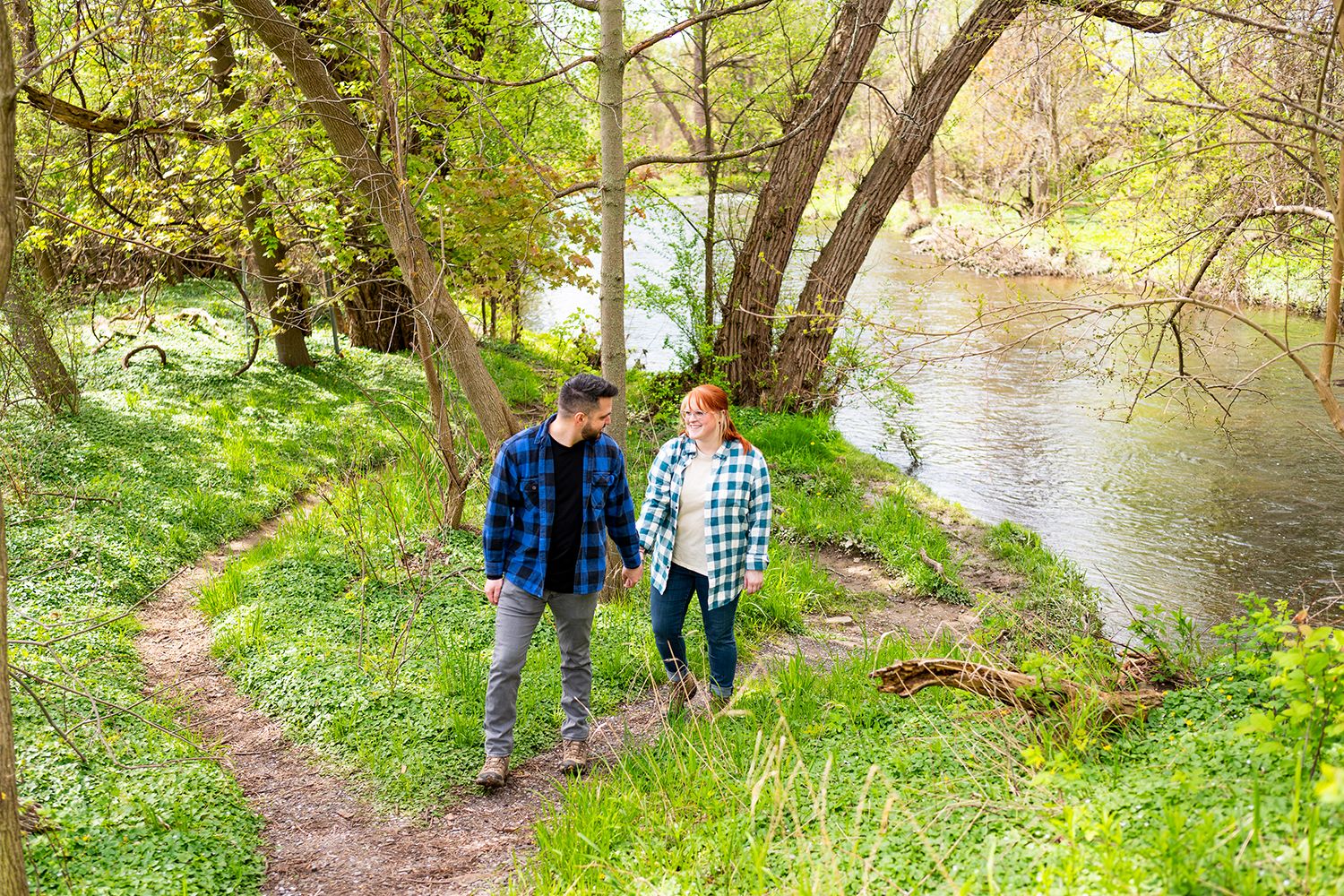 Hikers walking along a narrow dirt path through lush greenery beside a winding river.
