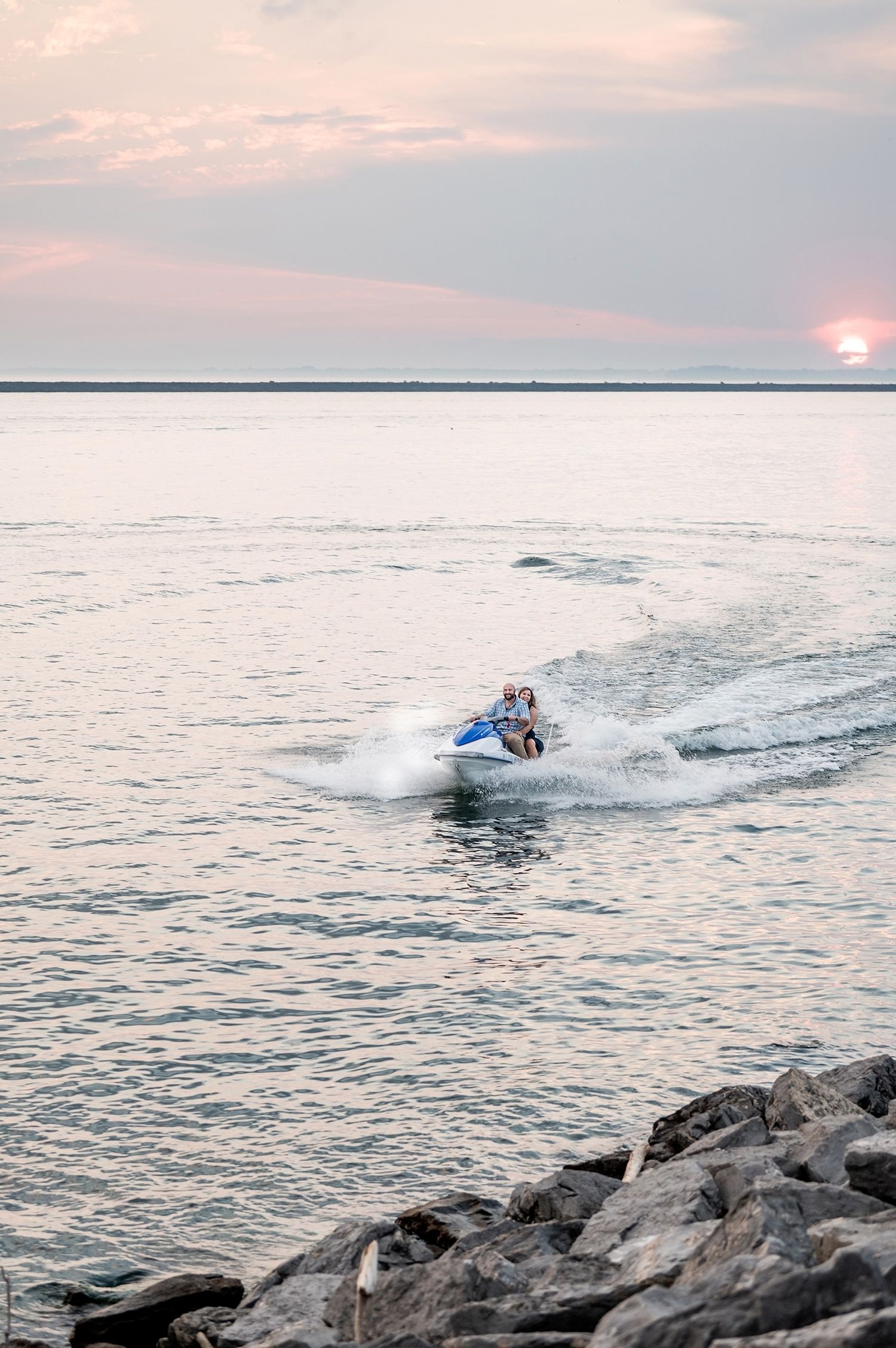 Two riders on a blue jet ski speed across calm ocean waters near rocky shore at sunset.