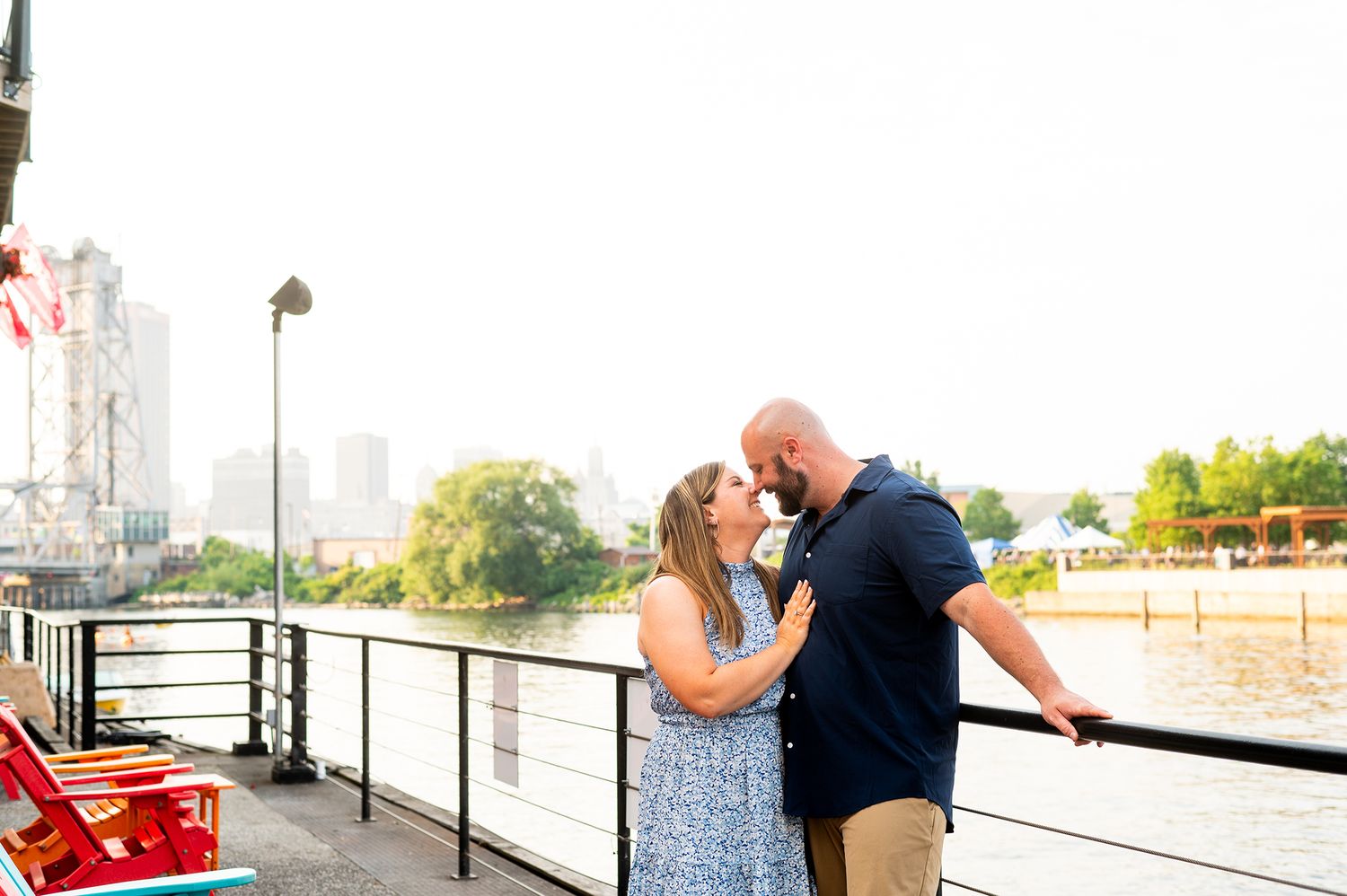 Couple sharing a romantic moment on a waterfront deck with a city skyline and river in the golden hour background.