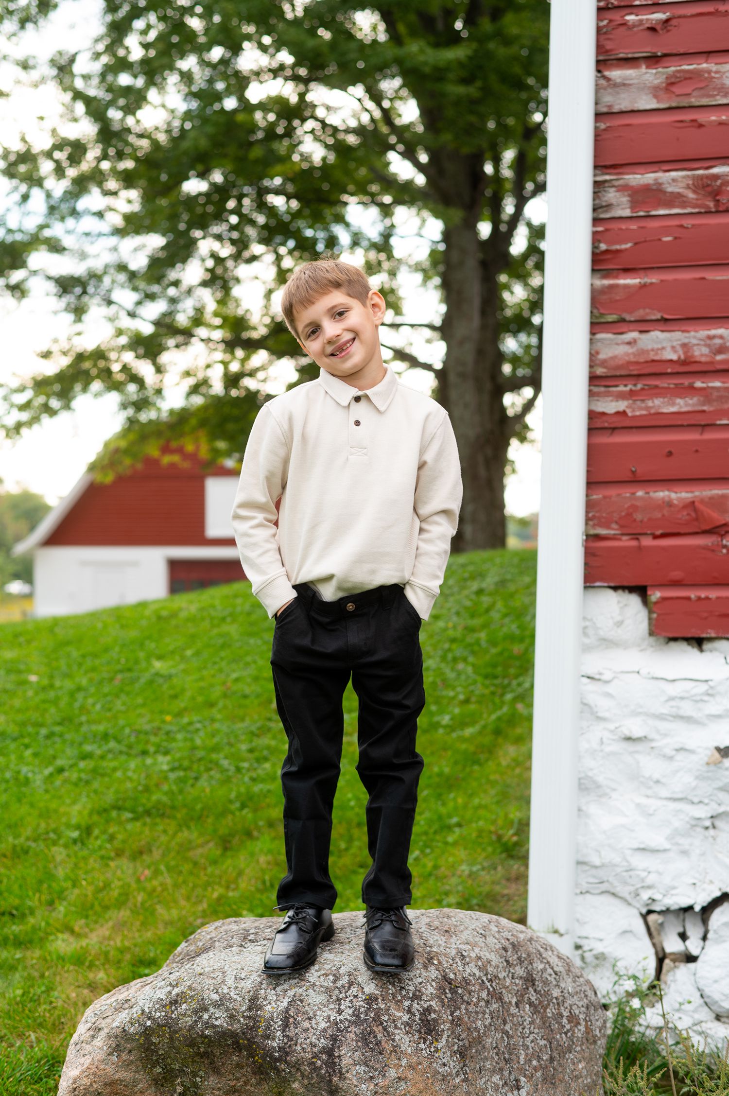 A smiling boy in a cream sweater and black pants stands on a rock near a red barn.