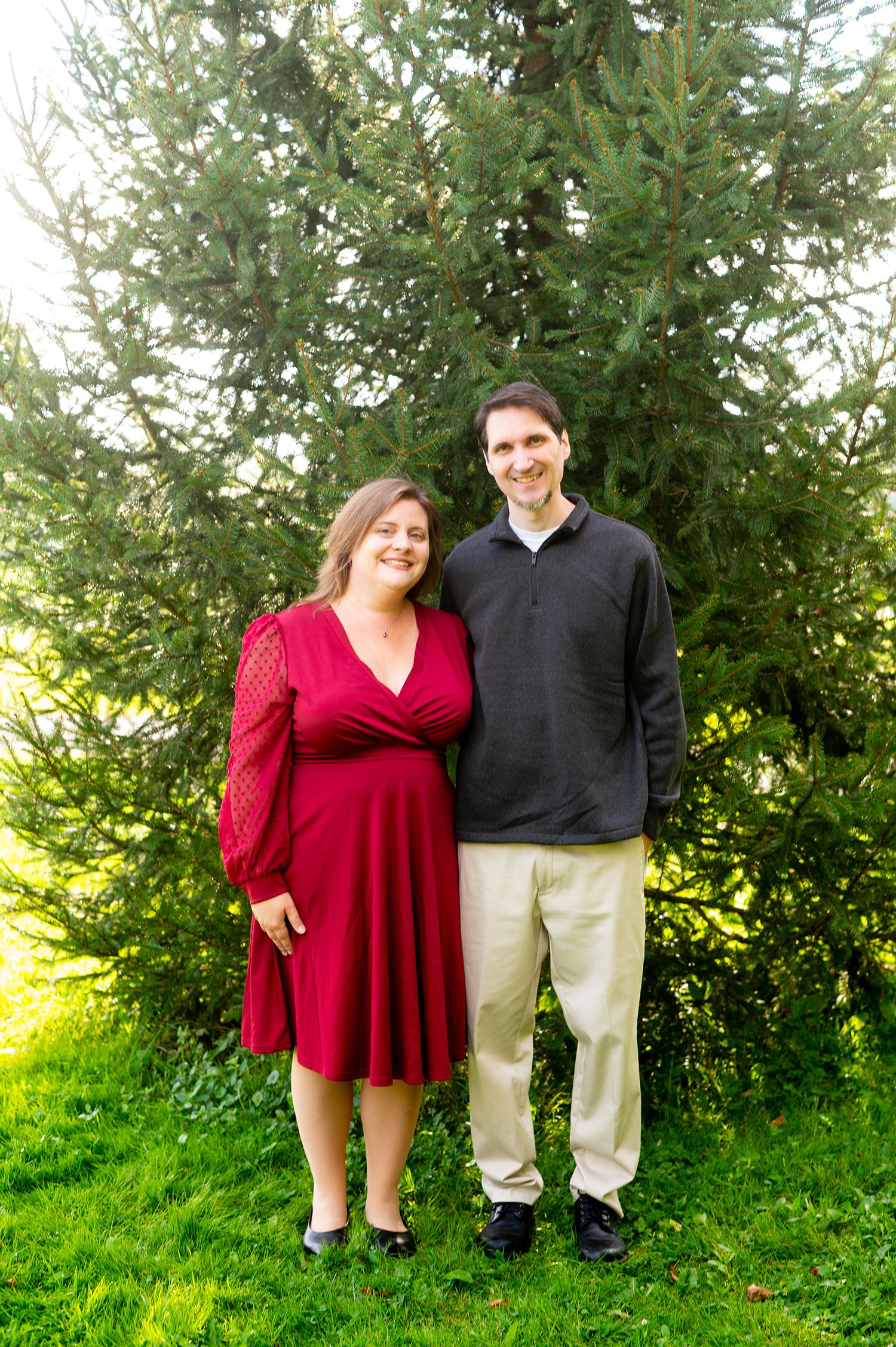 Smiling couple posing outdoors in front of evergreen trees, woman in red dress, man in dark pullover and khakis.