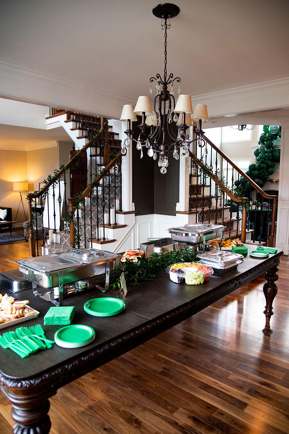 Elegant dining setup with green plates on dark wood table under crystal chandelier with staircase in background.