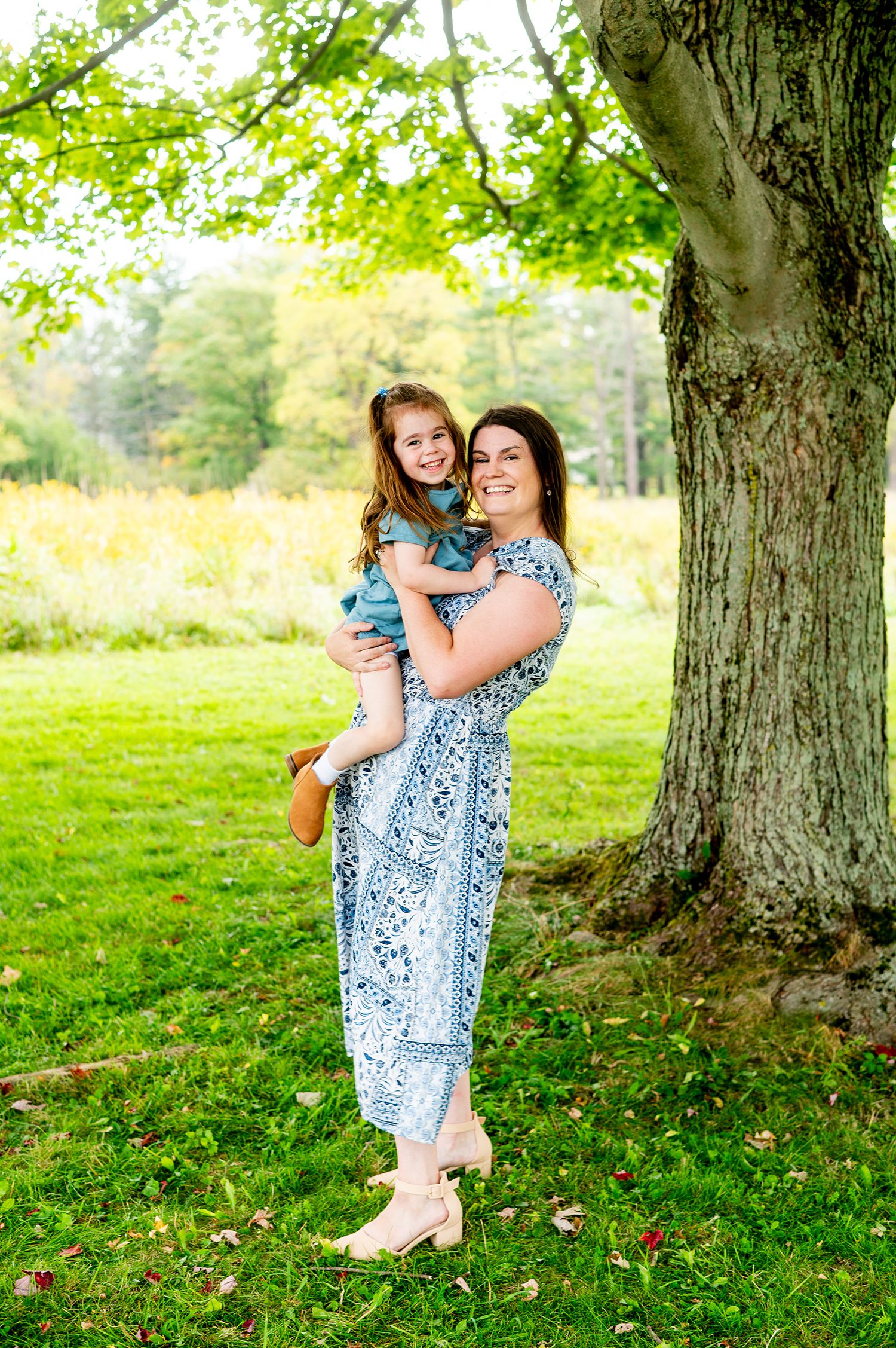 A smiling woman in a blue floral jumpsuit holds a young girl outdoors near a large tree on a lush green lawn.
