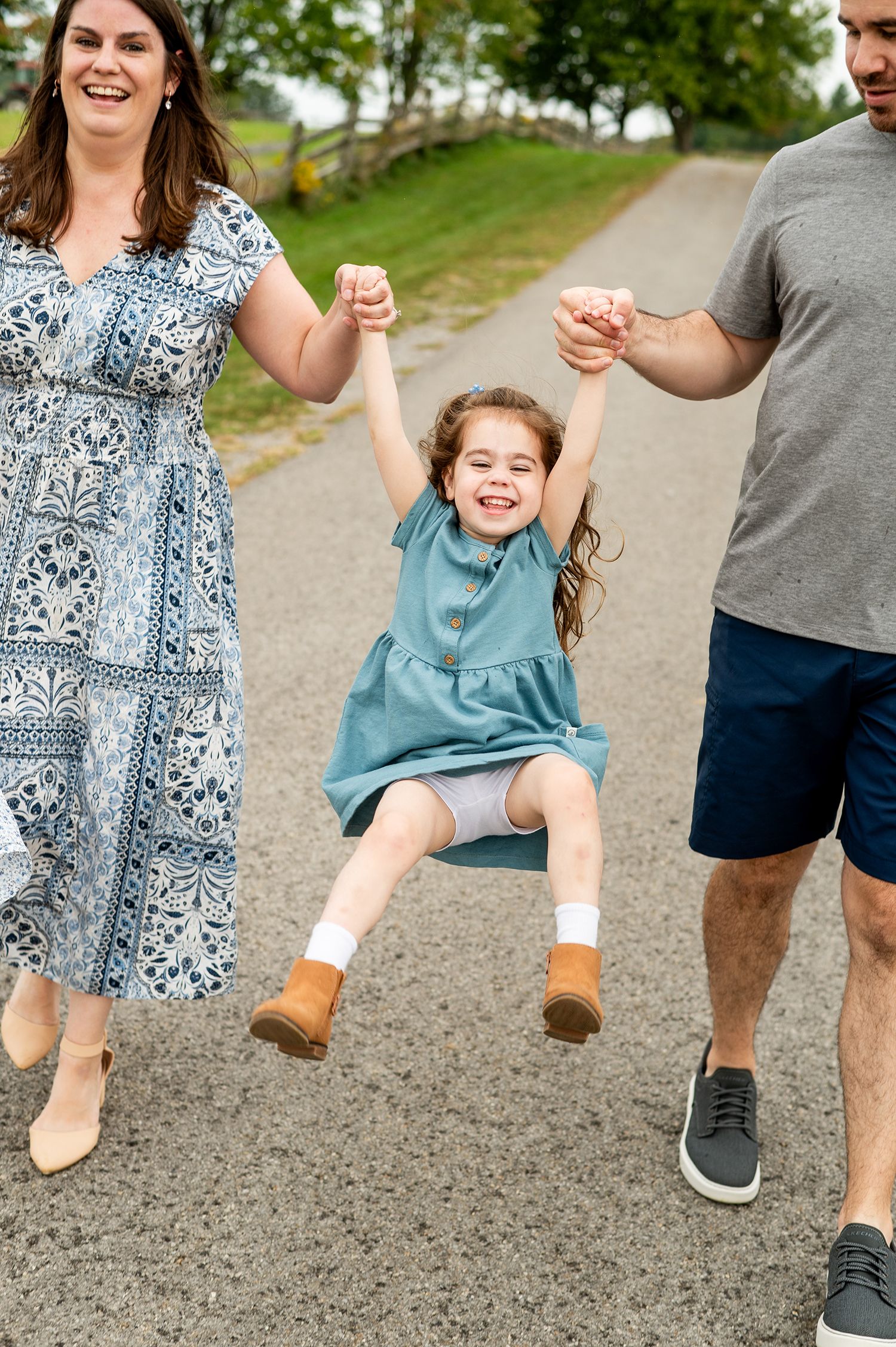 A joyful family swinging their little girl between them on a gravel path outdoors.