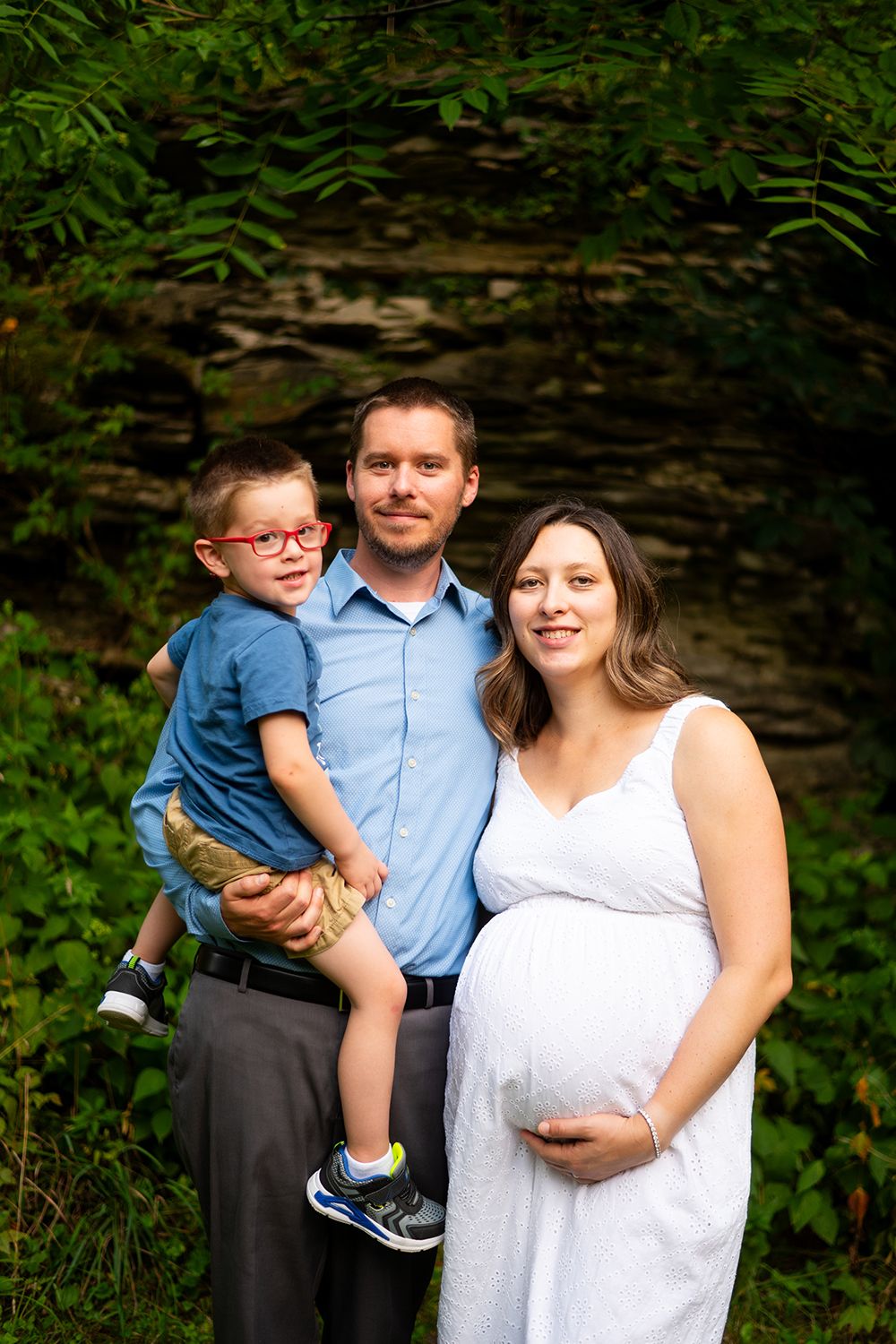 Family portrait in a natural outdoor setting with stone wall and green foliage.