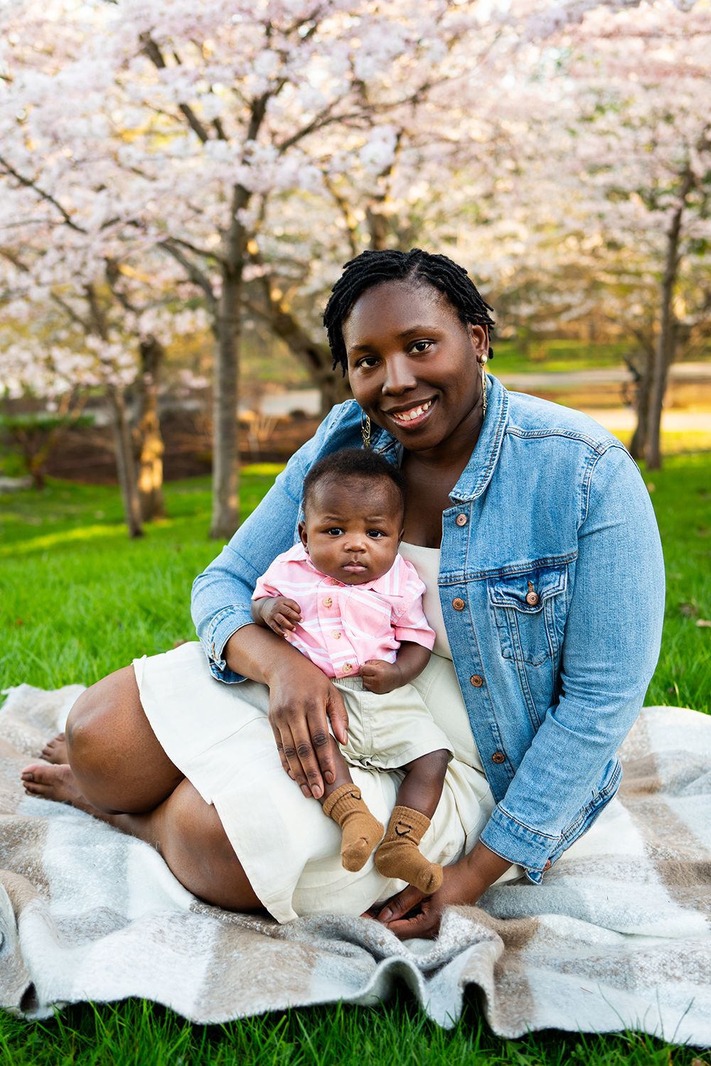 A parent and baby sit on a blanket under blooming cherry blossom trees on a sunny spring day.
