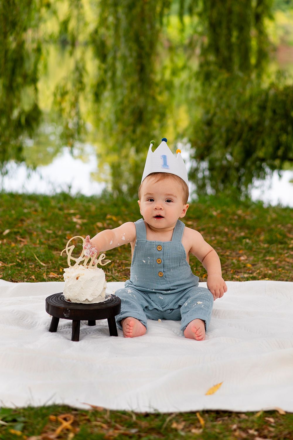 Small child in blue overalls sits on white blanket near drooping willow tree by a pond during cake smash photo session.