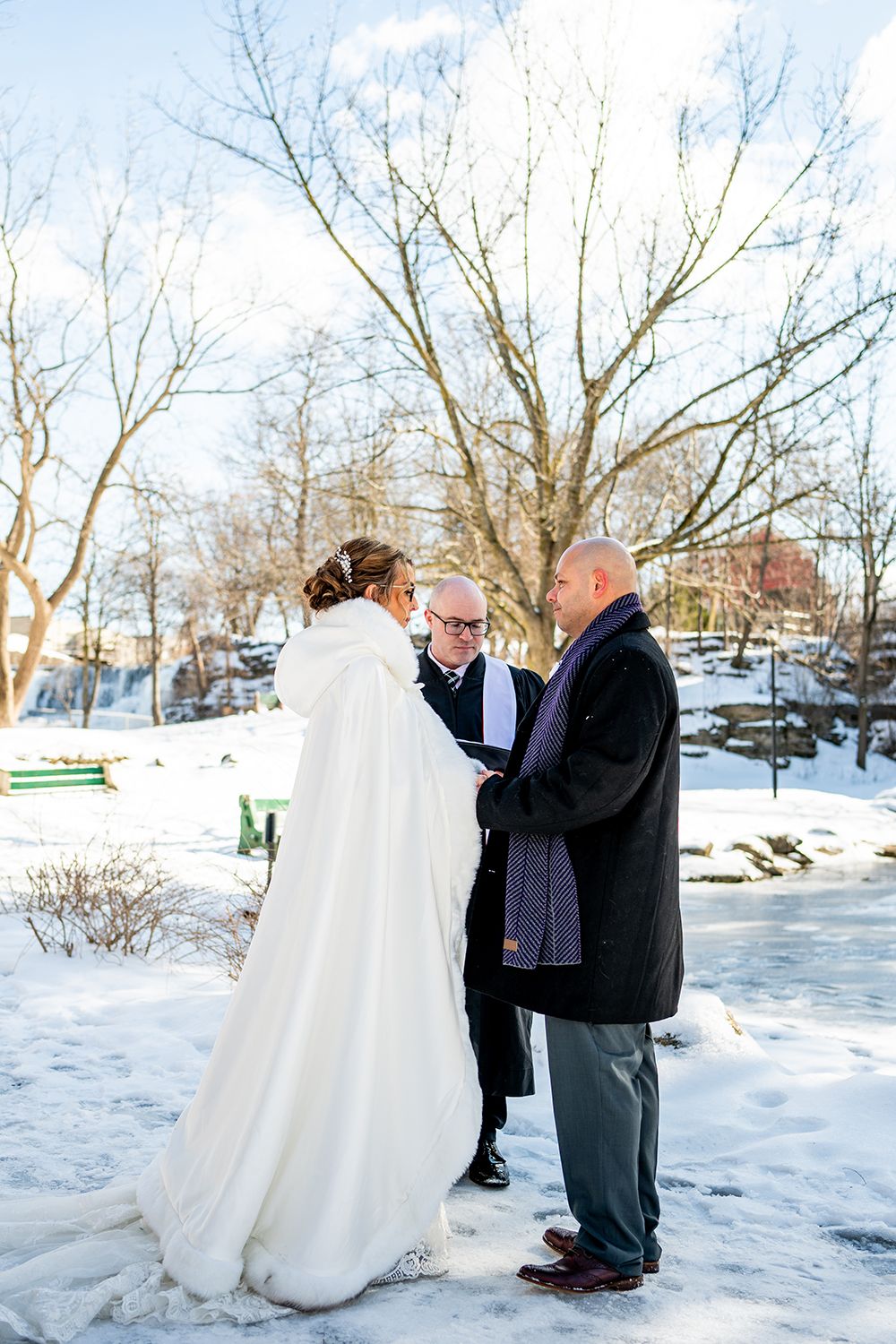 A winter wedding ceremony takes place outdoors in the snow with an officiant and couple wearing a long white cape.