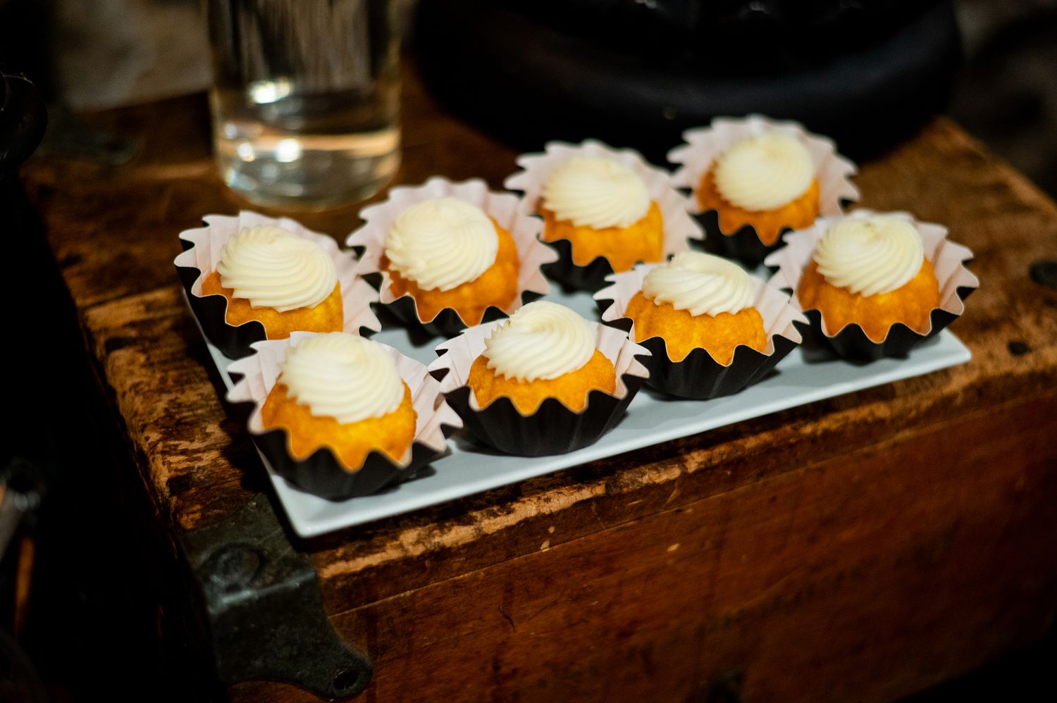Eight mini bundt cakes with white cream cheese frosting in black wrappers on a white rectangular tray.