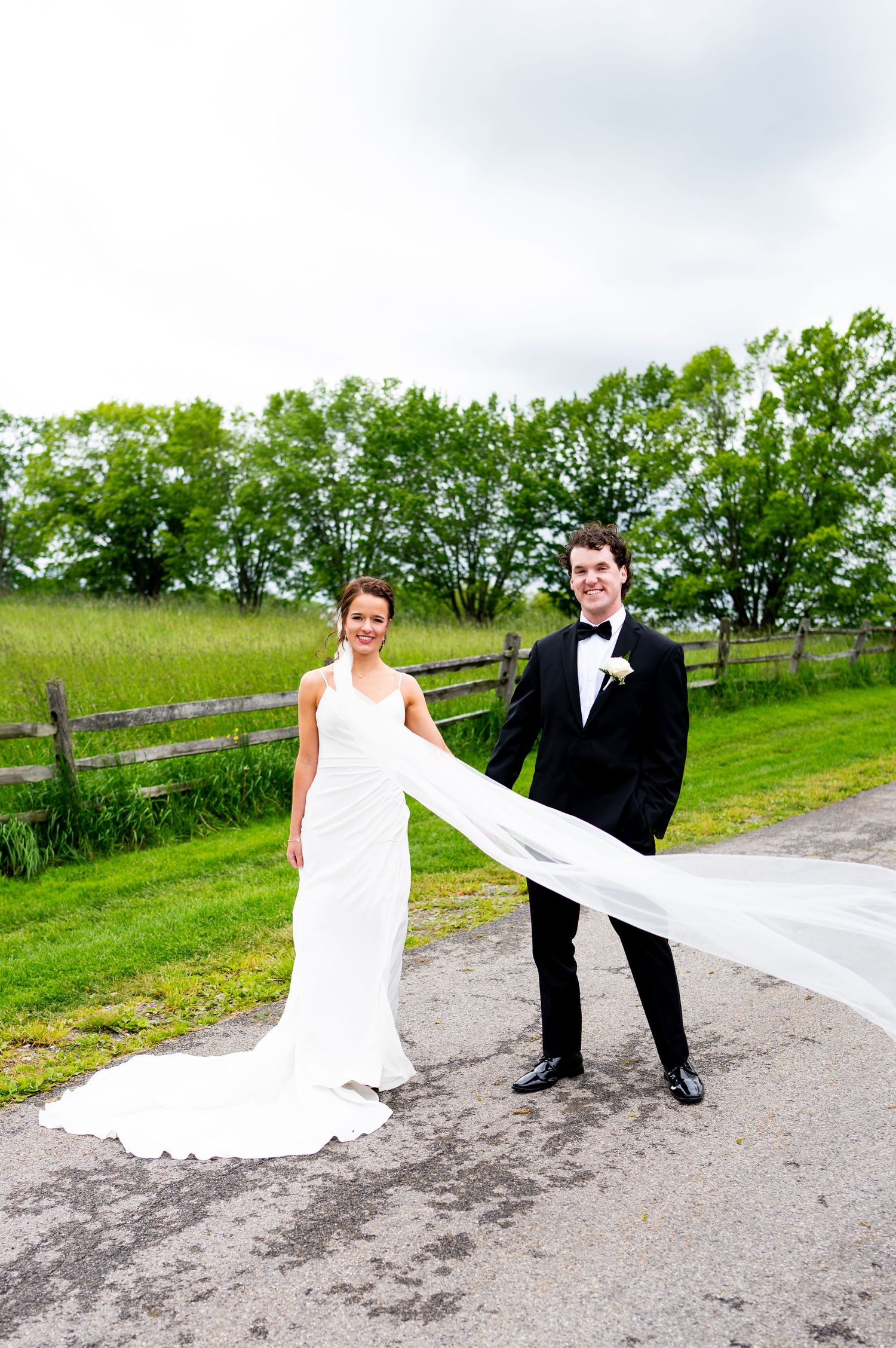 A bride and groom pose creatively with her flowing white dress train on a country road with green scenery.
