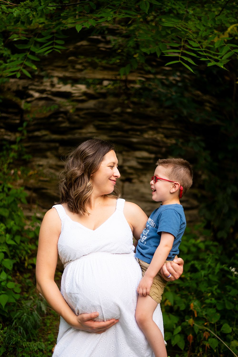 Outdoor maternity photo showing tender moment between woman in white dress and young child against rock wall background.