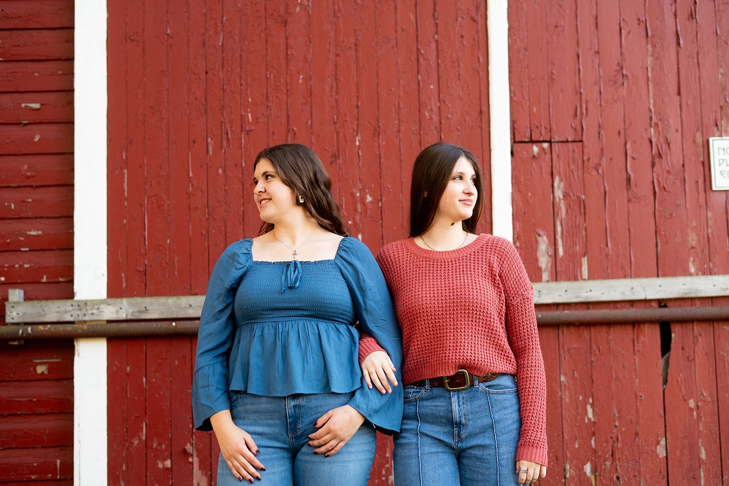 Two people wearing blue and rust-colored tops stand against a rustic red barn wall in a casual outdoor photo session.
