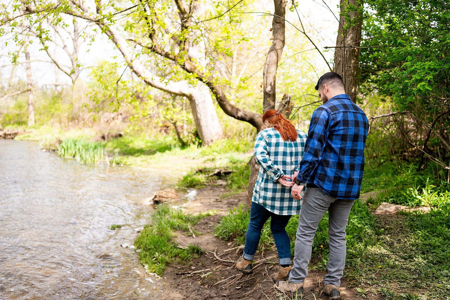 Two people wearing plaid shirts stand together by a peaceful river's edge on a sunny spring day.
