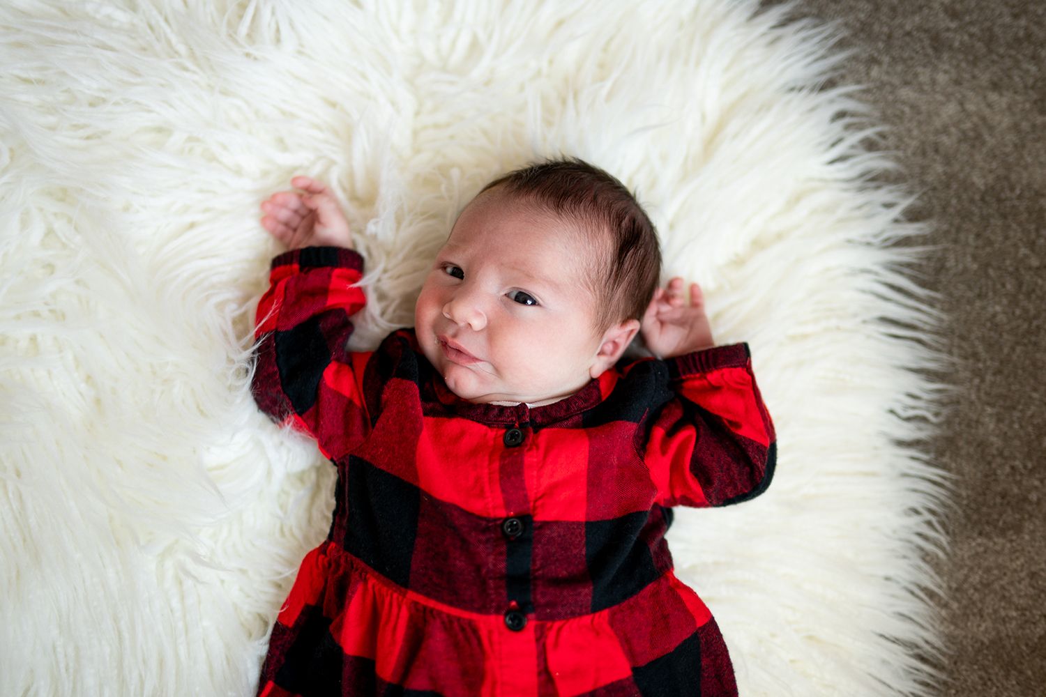 Baby wearing red and black buffalo plaid outfit lying on white fluffy rug with arms outstretched.