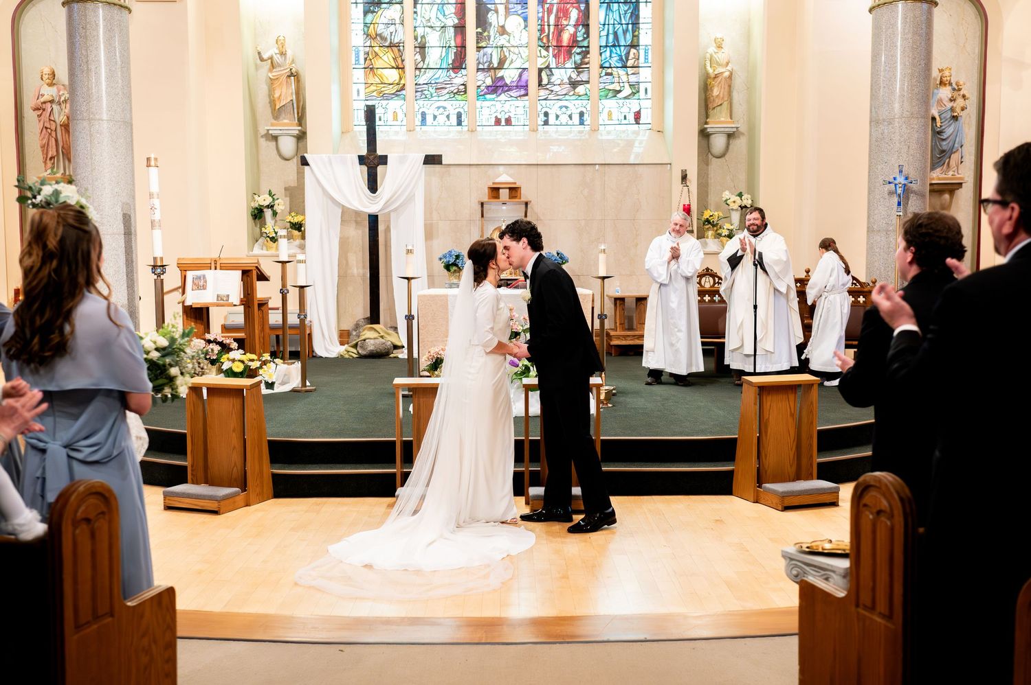 A wedding ceremony takes place in a beautiful church sanctuary with stained glass windows and traditional architecture.