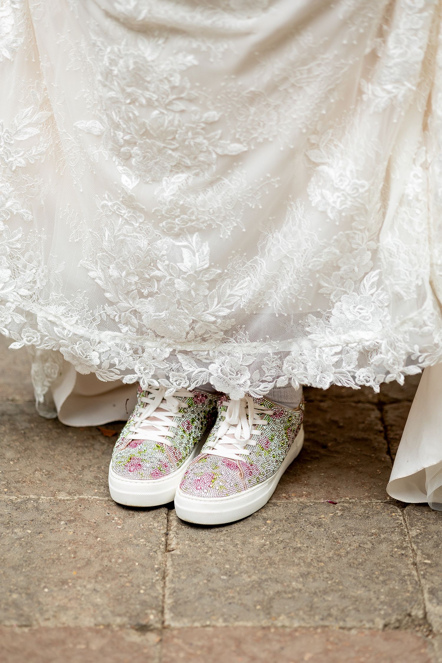 Bride wearing floral sneakers peeking beneath a white lace wedding gown on stone pavement.