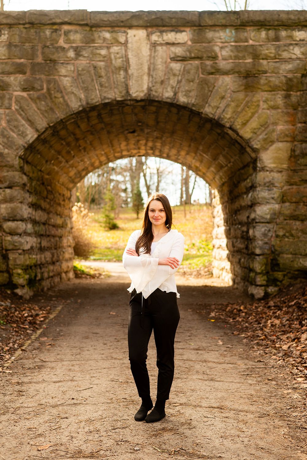 Figure stands in an arched stone tunnel walkway surrounded by autumn foliage with natural light filtering through.