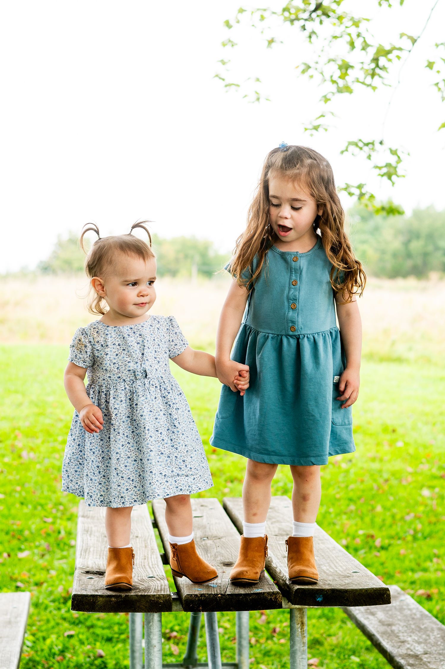 Two young girls in matching teal dresses and brown boots standing on a wooden picnic table outdoors.