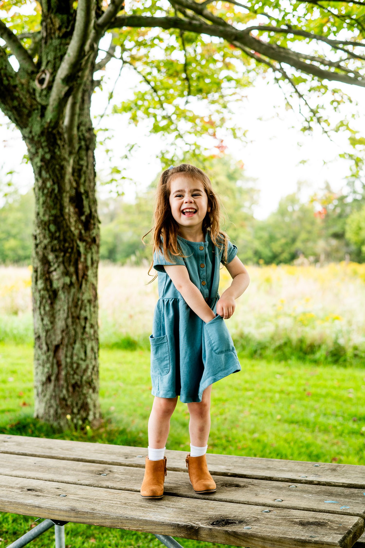 Young girl in teal dress and brown boots laughing while standing on a wooden picnic table outdoors.