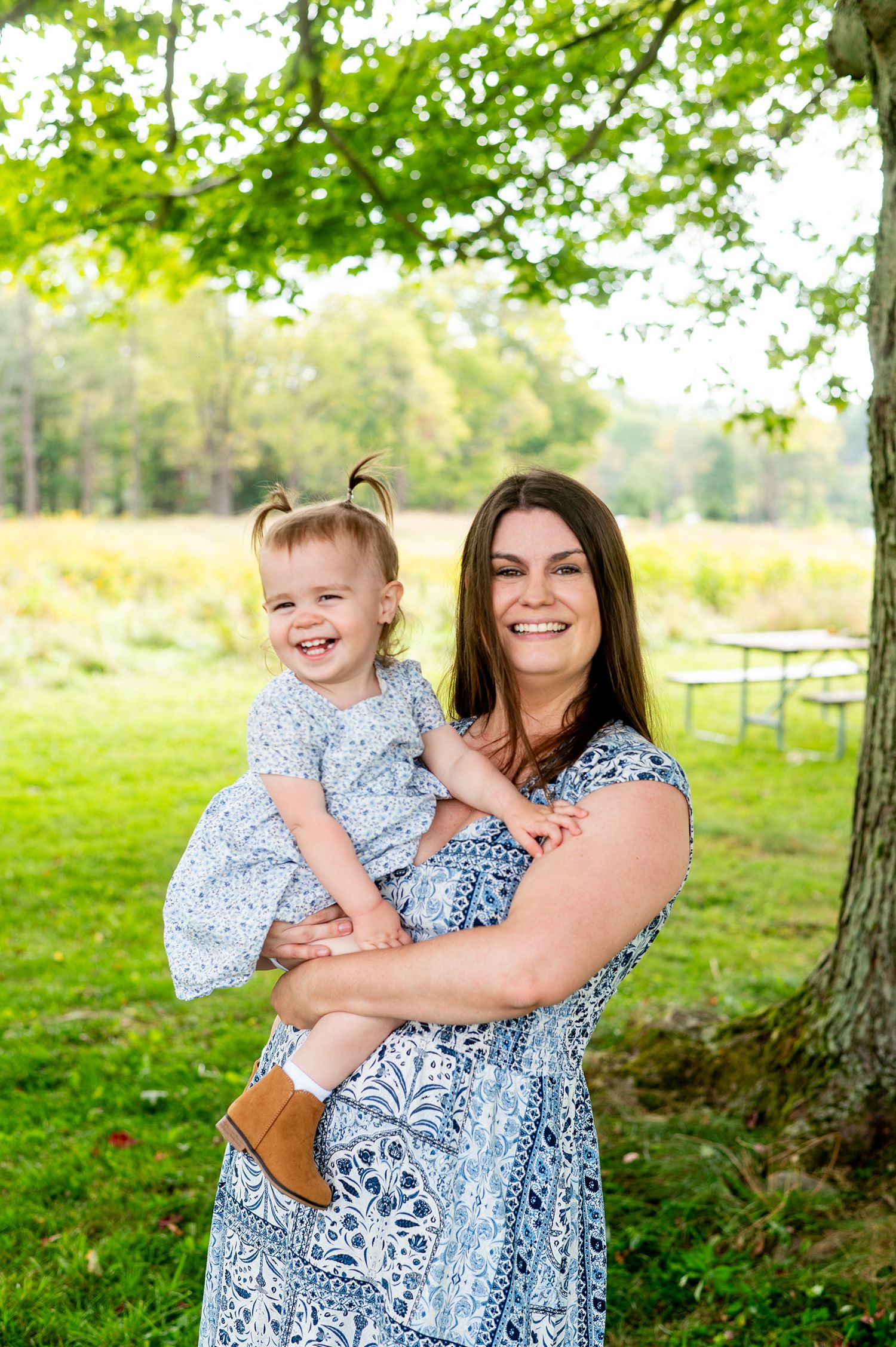 A smiling woman in a blue floral dress holds a laughing toddler girl outdoors under a green tree.