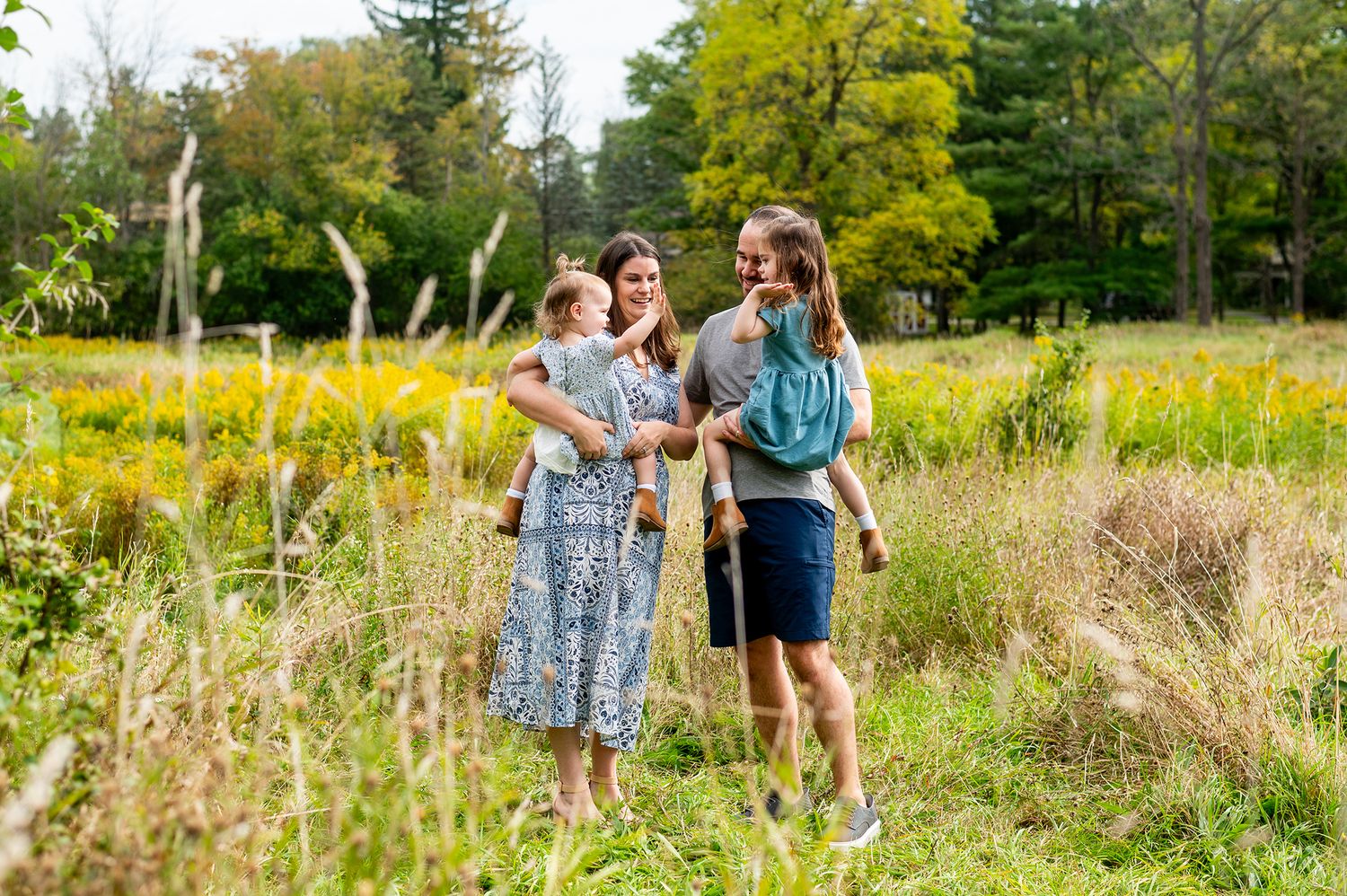 Family of four walking through a lush green meadow, parents carrying young children, smiling outdoors in nature.