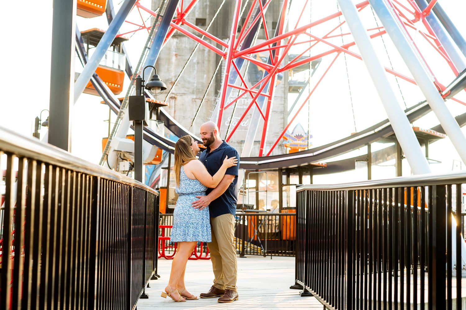 Couple embracing romantically in front of a colorful Ferris wheel at an amusement park on a sunny day.