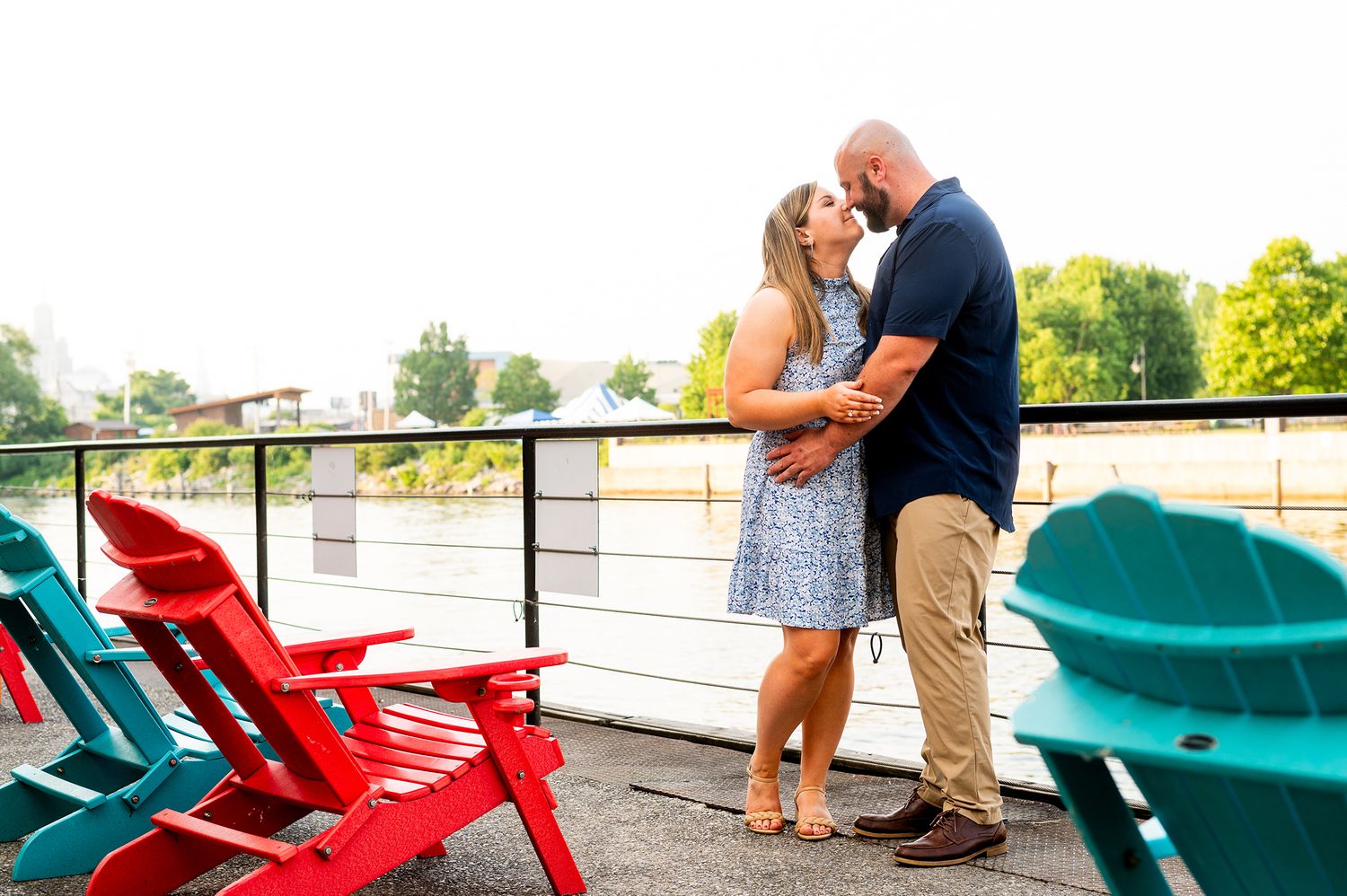 Couple kissing on a waterfront deck surrounded by colorful Adirondack chairs in red and teal.