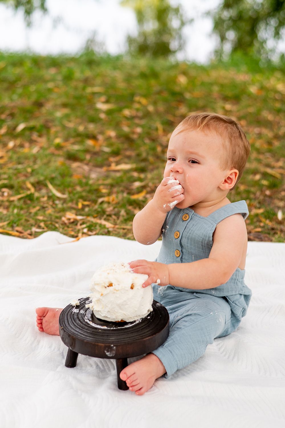 Baby in blue overalls sits on white blanket eating cake from a black plate on grass and fallen leaves.