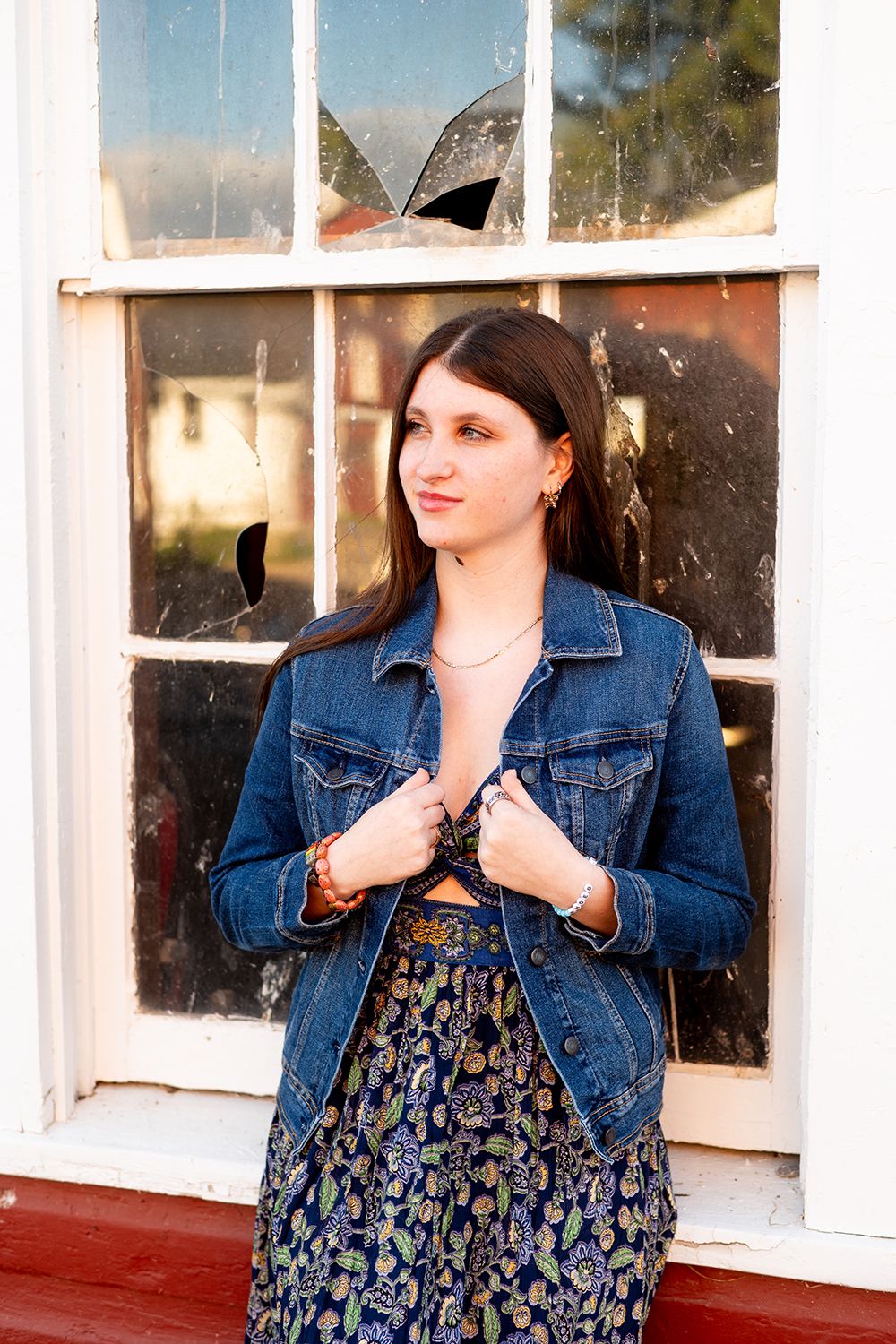 A person stands by a window wearing a denim jacket and floral dress in a casual outdoor portrait setting.