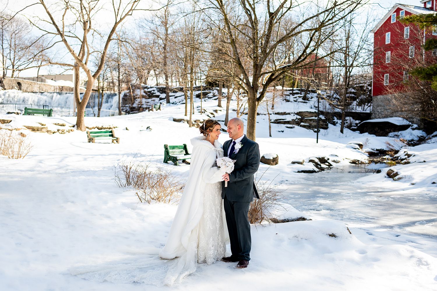 A couple shares an intimate winter wedding moment in a snowy park with a rustic red building in the background.