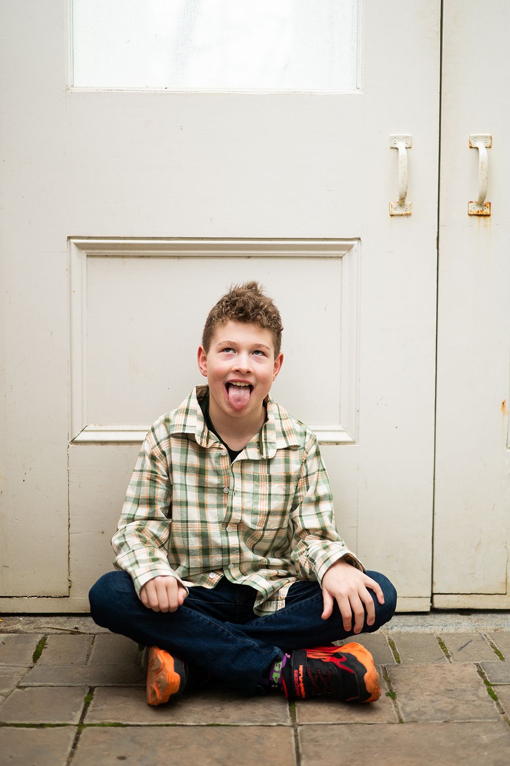 A playful cross-legged pose against a gray door while wearing a plaid shirt and jeans.