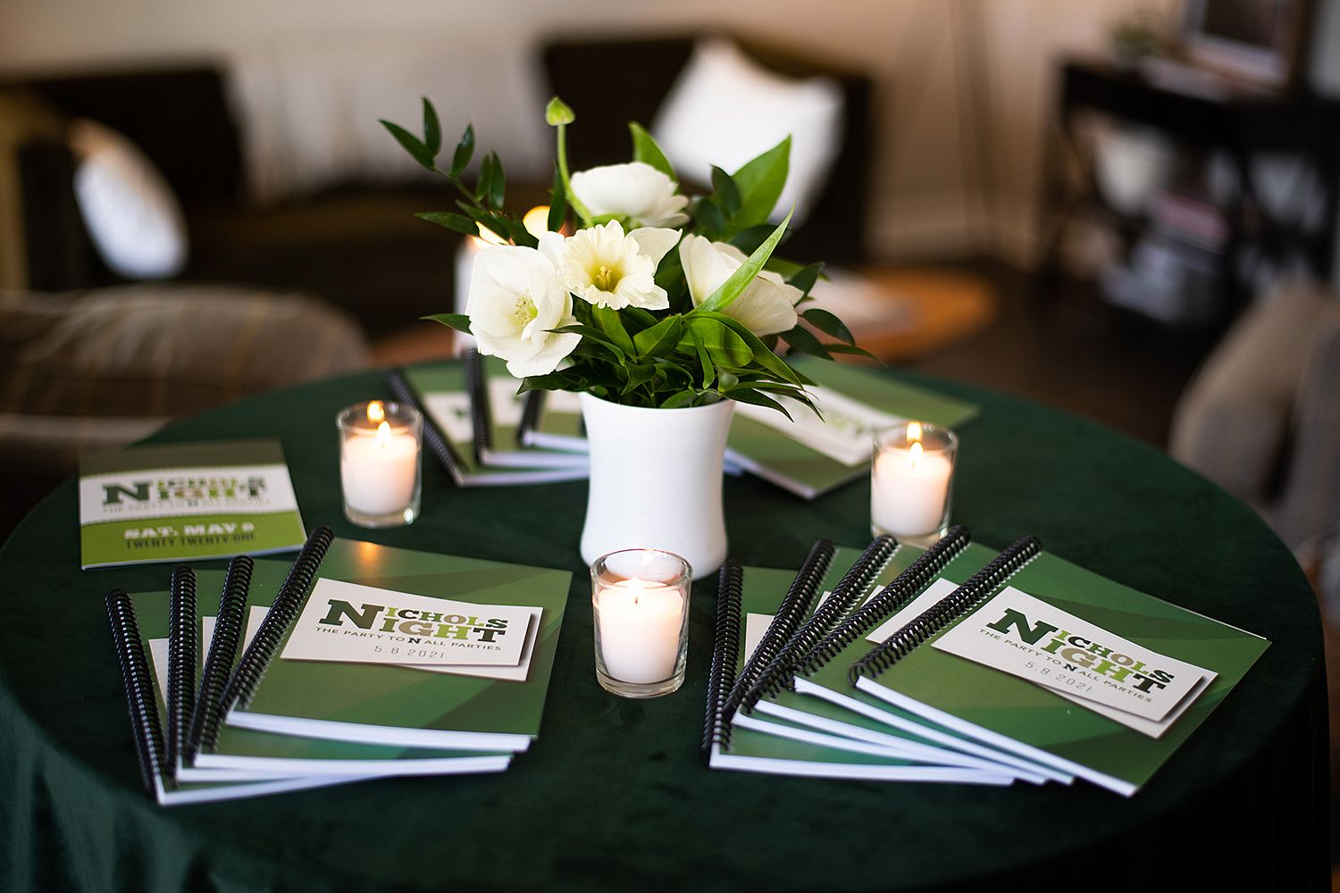 White floral centerpiece in vase with candles and stacked programs on dark green tablecloth at elegant event.