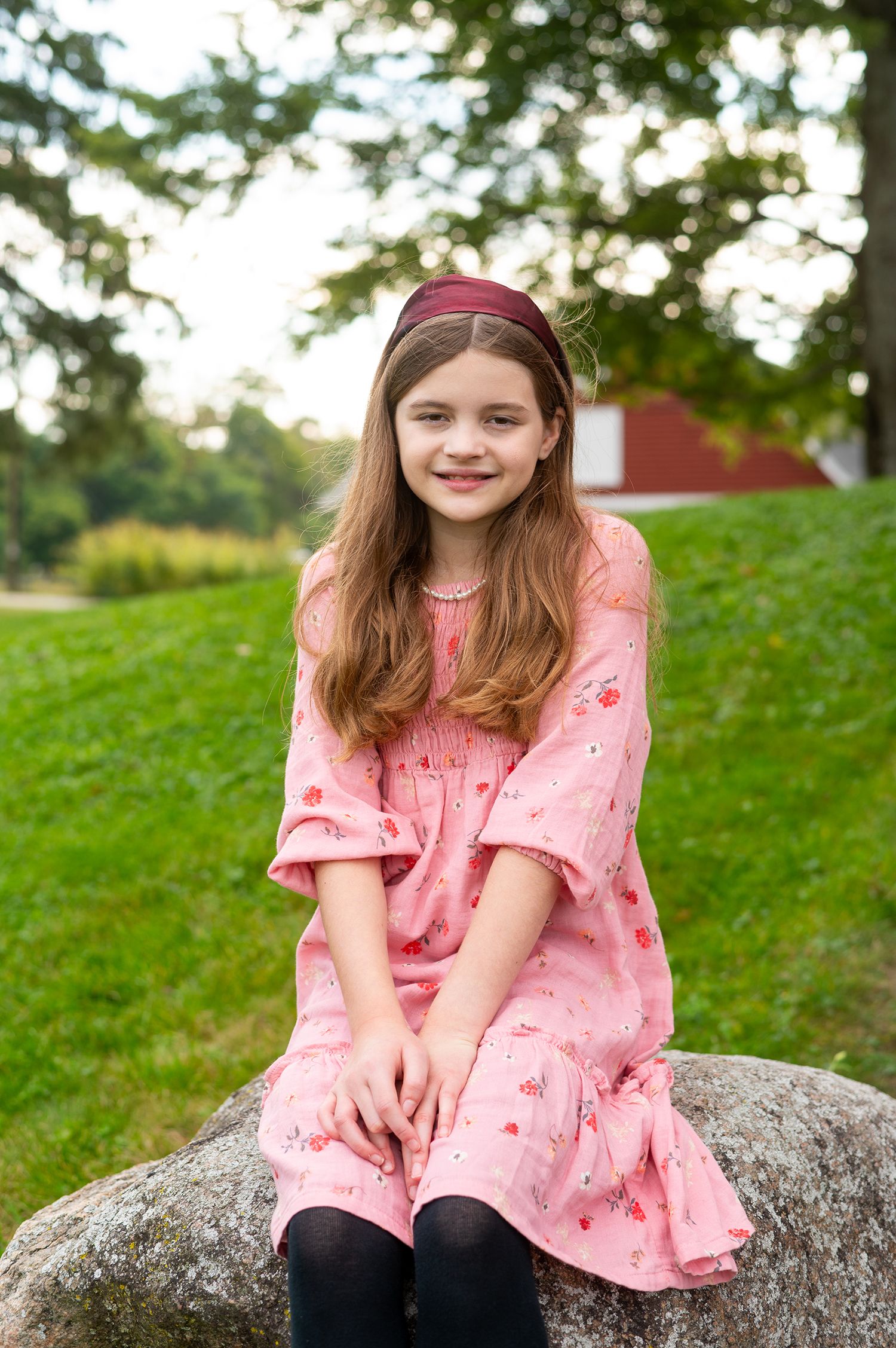 A young girl in a pink floral dress and burgundy headband sits smiling on a rock outdoors.