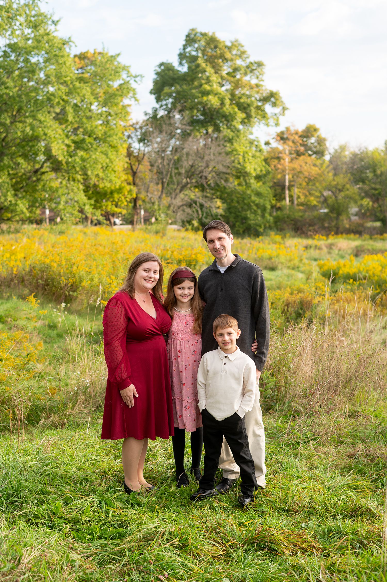 Family of four smiling outdoors in a green meadow with yellow wildflowers, wearing coordinated outfits in fall.