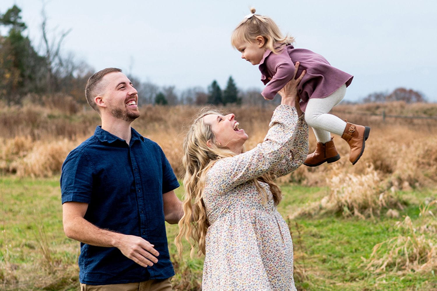 A joyful family outdoors, mom lifting toddler girl in the air while dad smiles, in a scenic field setting.