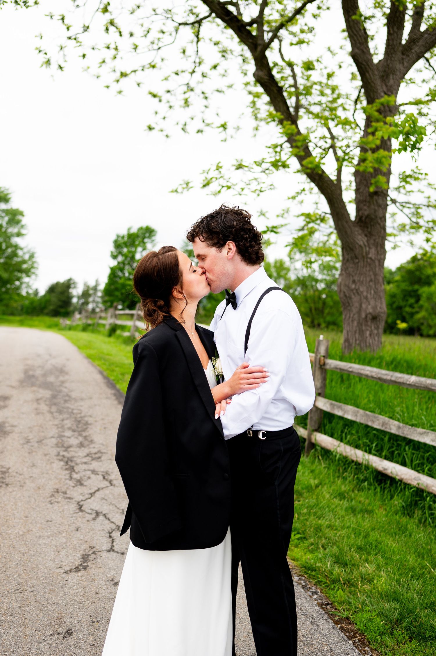 A romantic couple shares tender moments along a tree-lined path with a rustic wooden fence on a spring day.