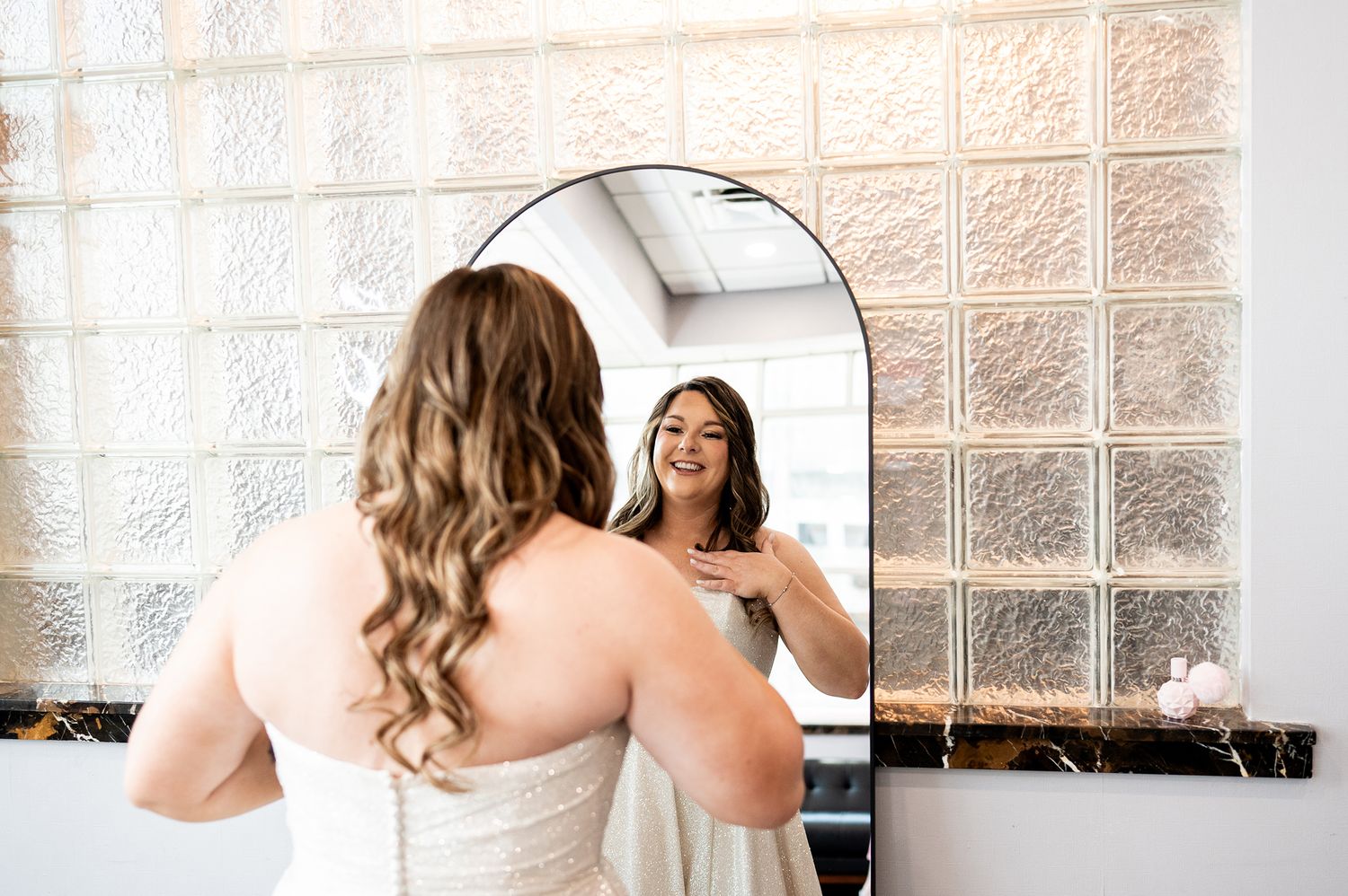 A bride in a strapless white wedding gown stands in front of a window with textured glass blocks.