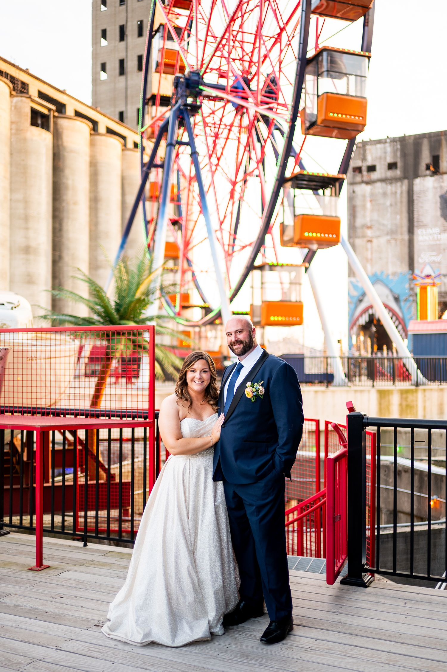 A couple poses together on a boardwalk with a red carnival ride structure in the background.