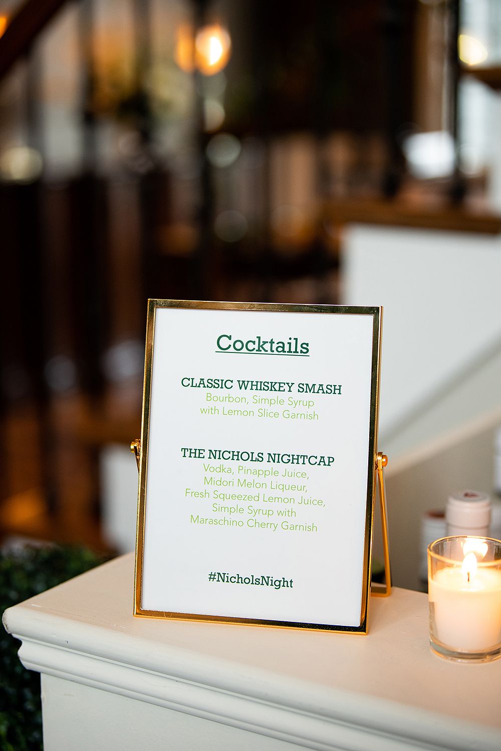 A framed cocktail menu displays drink options including Classic Whiskey Smash next to a glowing white candle in a bar setting.