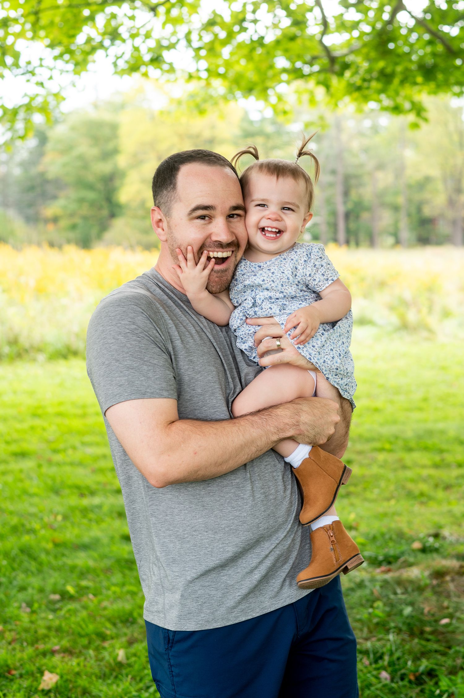 A joyful father holds his laughing toddler girl outdoors in a lush green park setting.