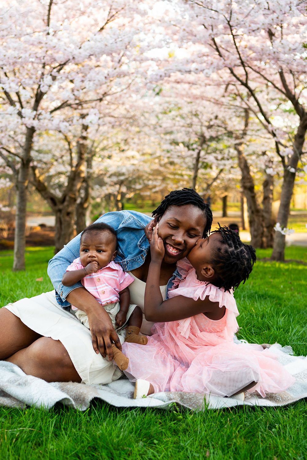A family plays and cuddles together on a blanket under blooming cherry blossom trees in a spring park.