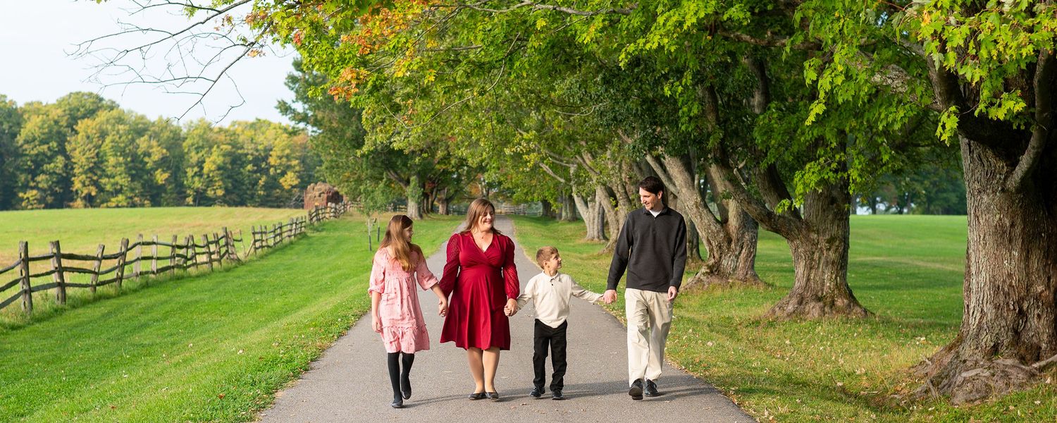 Family of four walking together on a tree-lined path through a lush green park on a sunny autumn day.