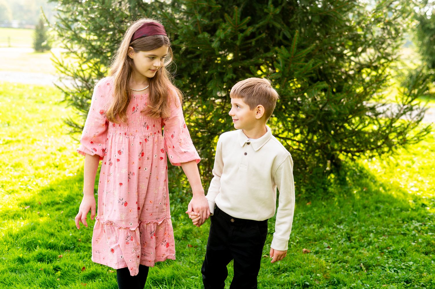 Two children holding hands in a sunny green park, girl in pink dress and boy in cream sweater walking together.