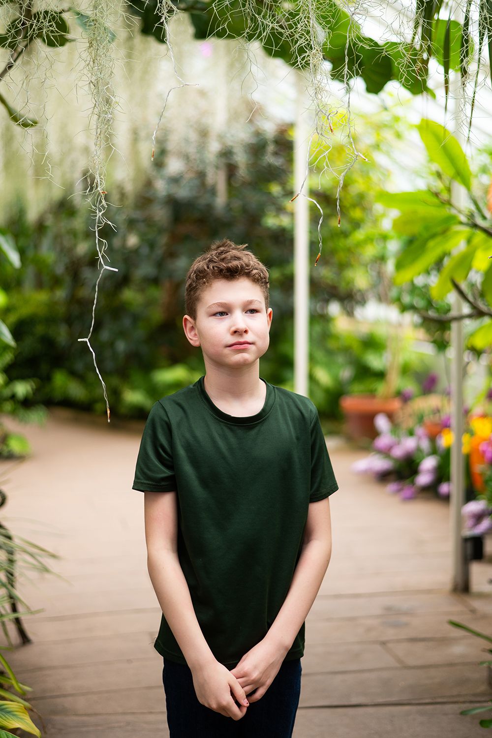A figure in a dark green t-shirt stands in a lush greenhouse setting surrounded by tropical plants and flowers.