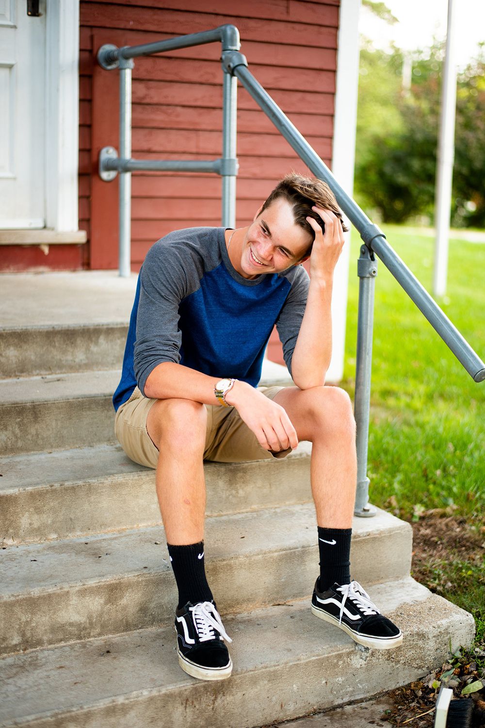 Someone sits on concrete steps beside a red building wearing a blue shirt, khaki shorts and black sneakers.