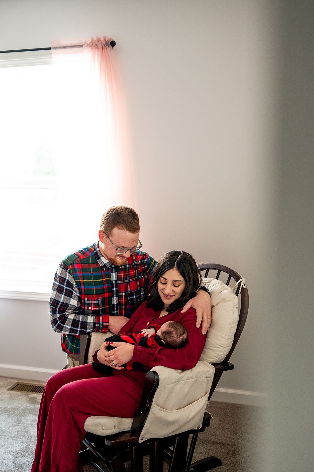 A couple sits together in a white rocking chair while holding a baby wearing red clothing.