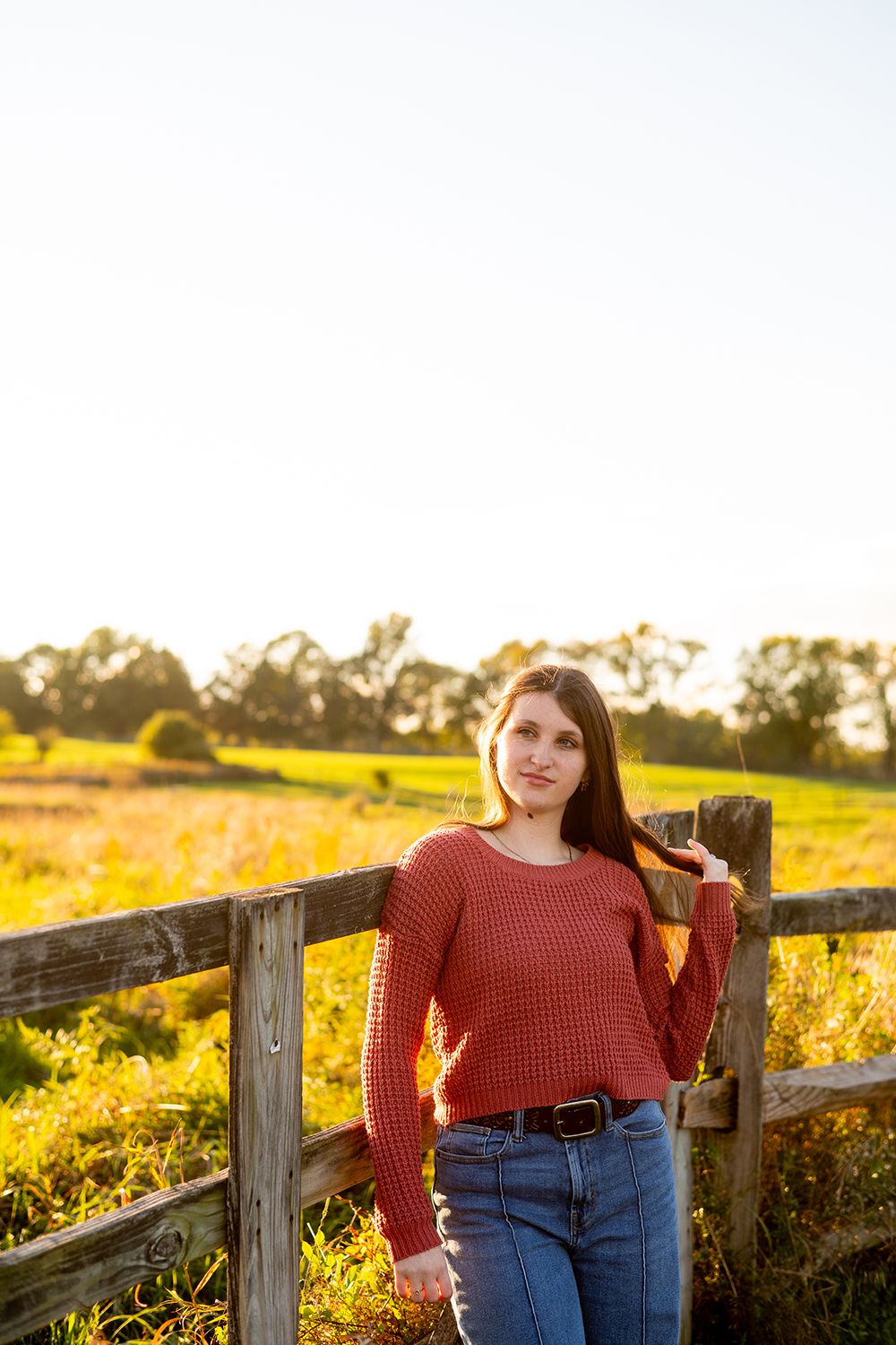 A figure in a red sweater and jeans poses by a wooden fence at golden hour in a rural field.