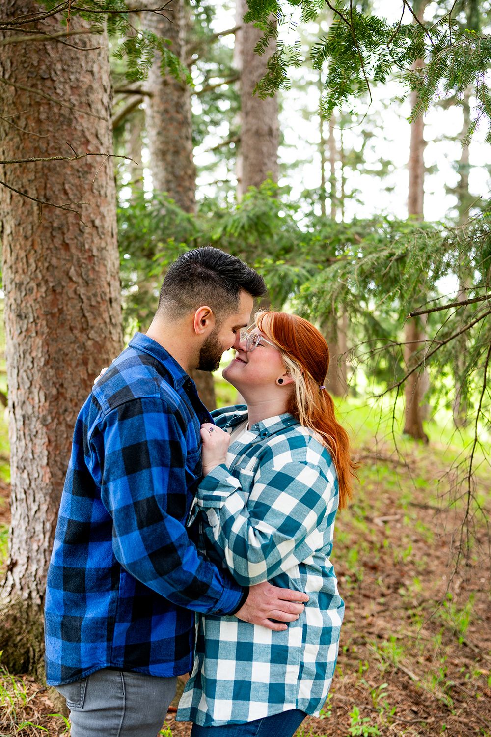 A couple wearing matching plaid flannel shirts embraces romantically in a sunlit pine forest setting.