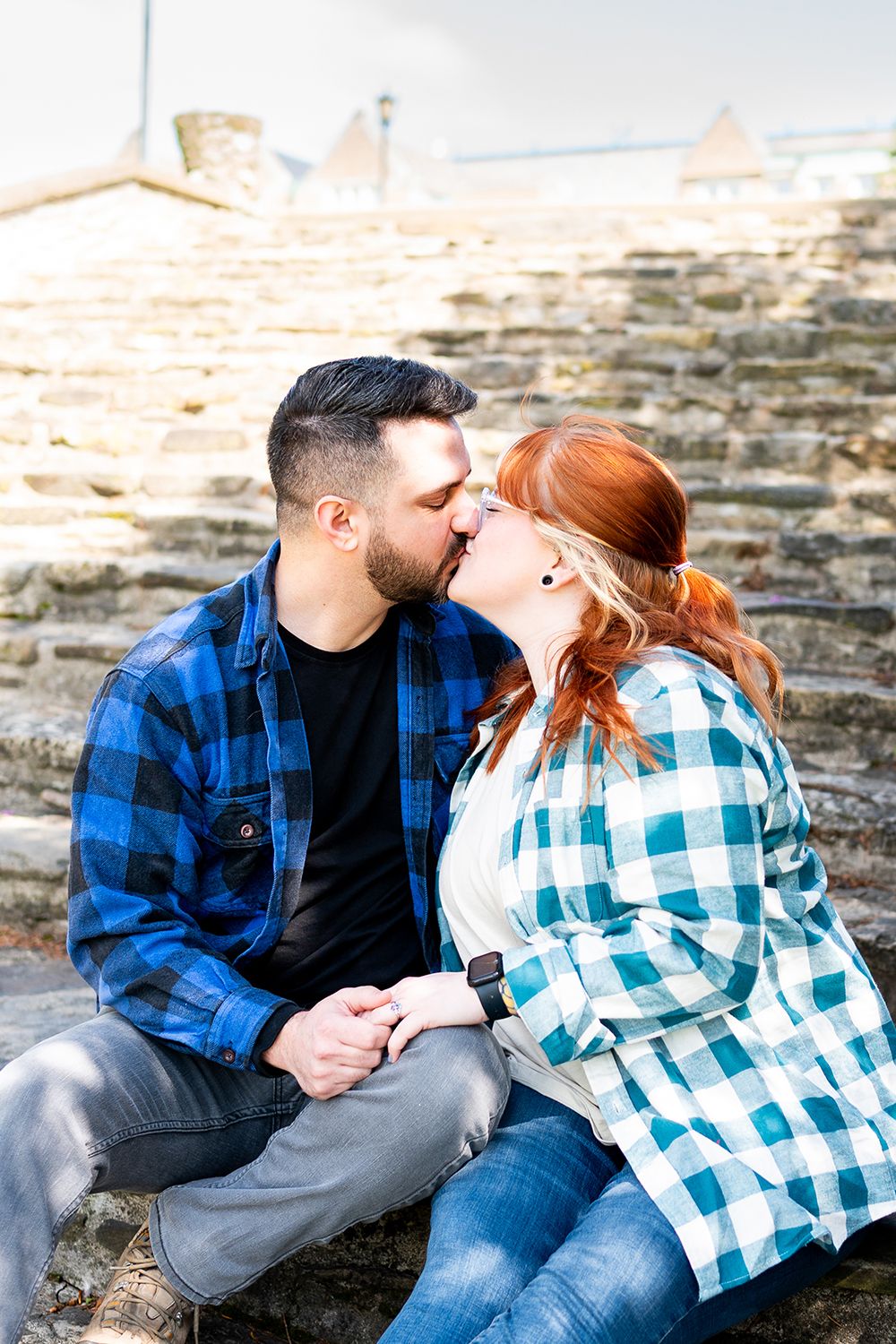 A couple wearing plaid and casual attire share romantic moments while sitting on stone steps outdoors.